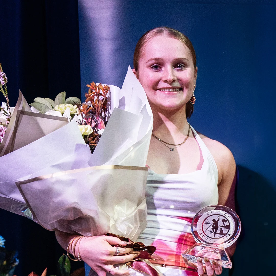 A smiling young woman onstage holds a large bouquet and a clear award, wearing a white top and pink sash against a blue backdrop; celebratory, proud mood.