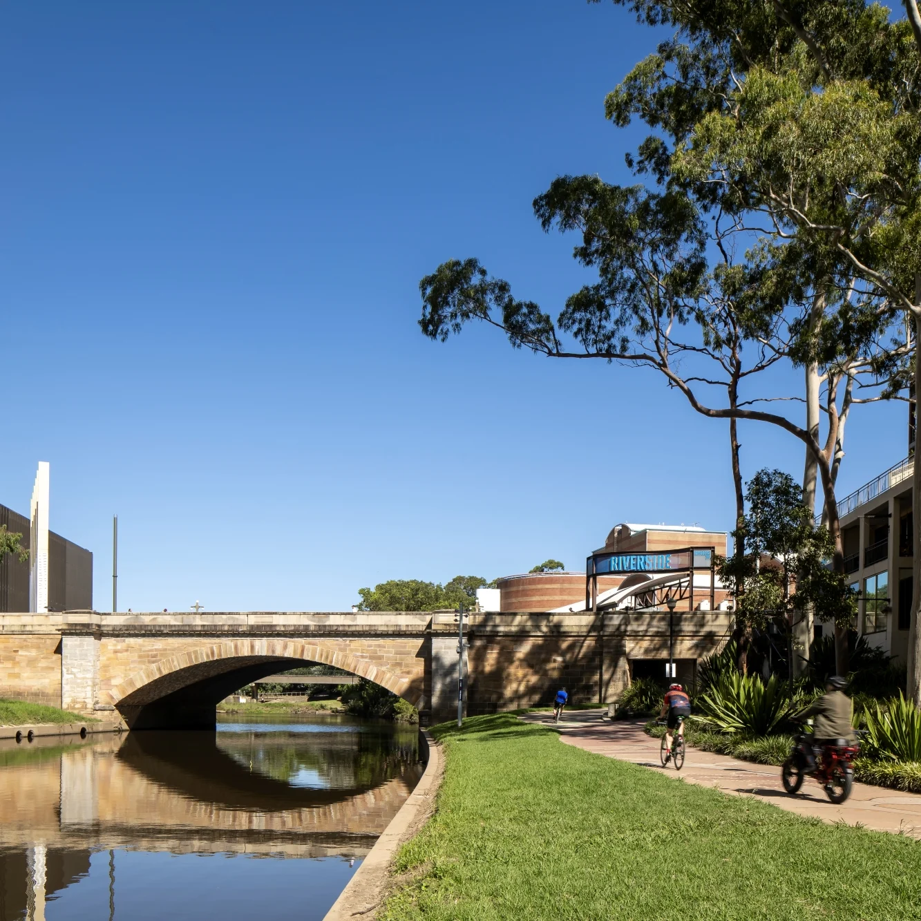 A sunny urban riverside scene with a stone bridge reflected in calm water, high-rise buildings, leafy trees, and a cyclist on the path; peaceful and open.