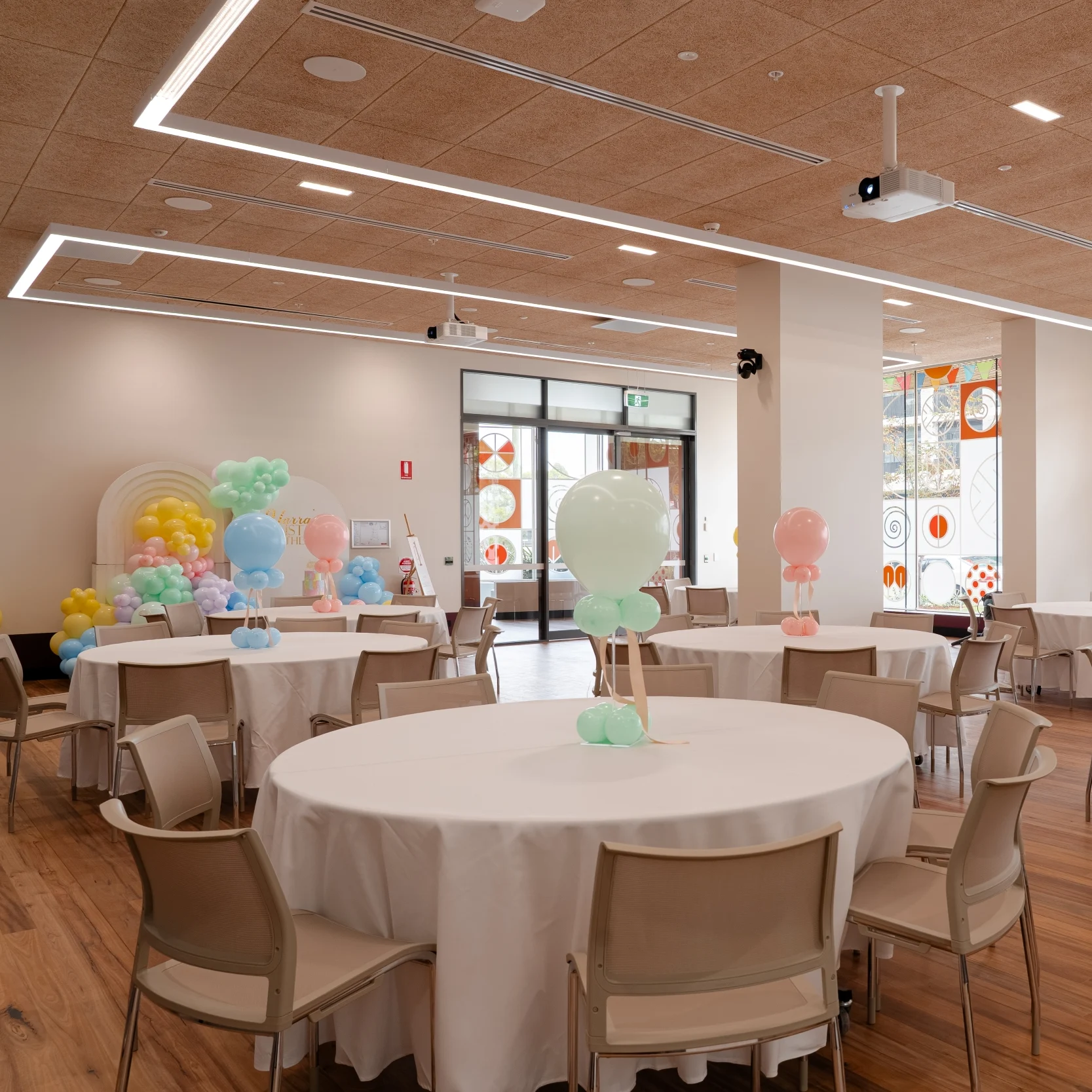 A bright banquet room features round tables with white cloths, beige chairs, colourful balloons, and patterned windows, creating a cheerful party setting.