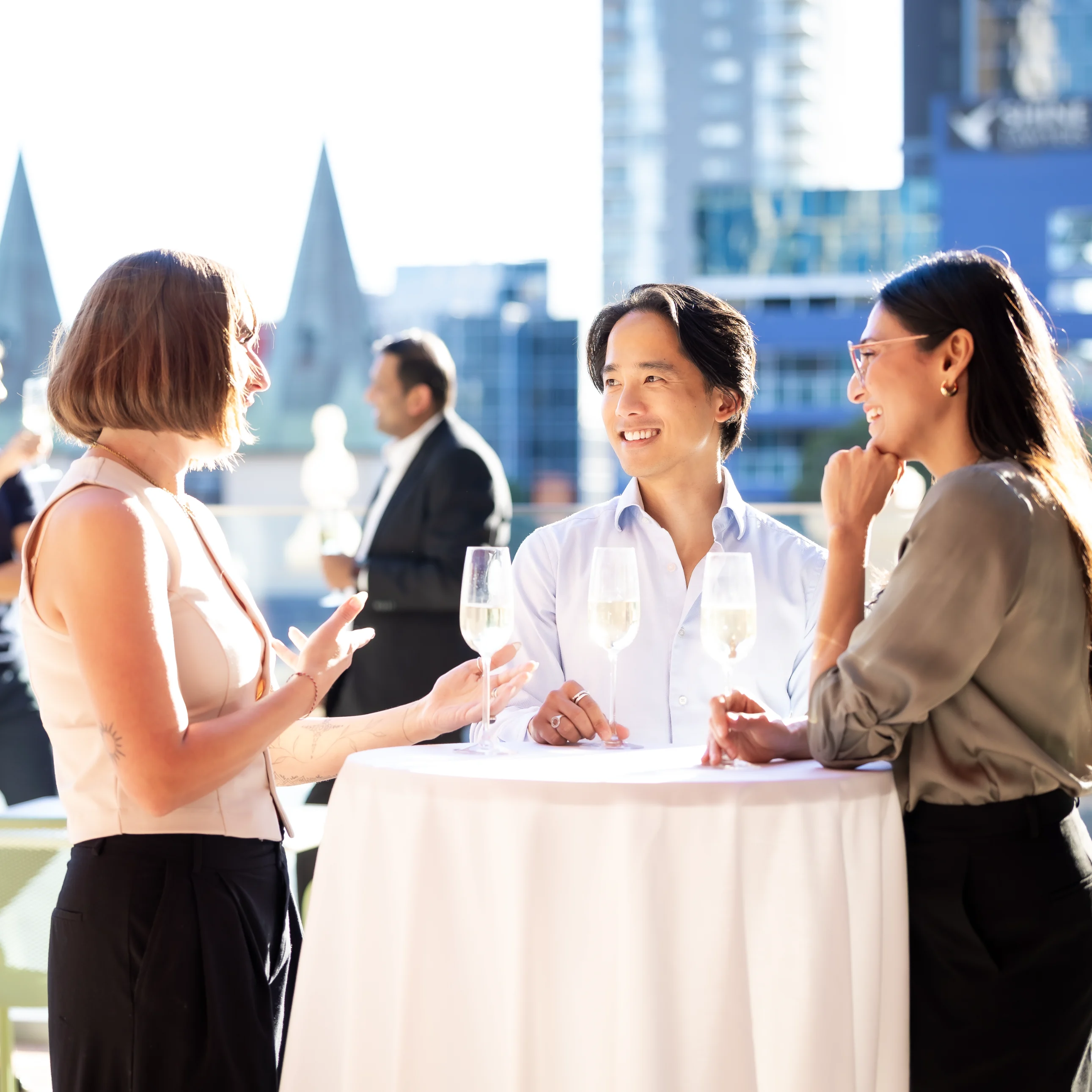 At a rooftop networking reception, three colleagues chat around a white cocktail table as guests mingle against a bright city skyline; the mood is relaxed and professional.