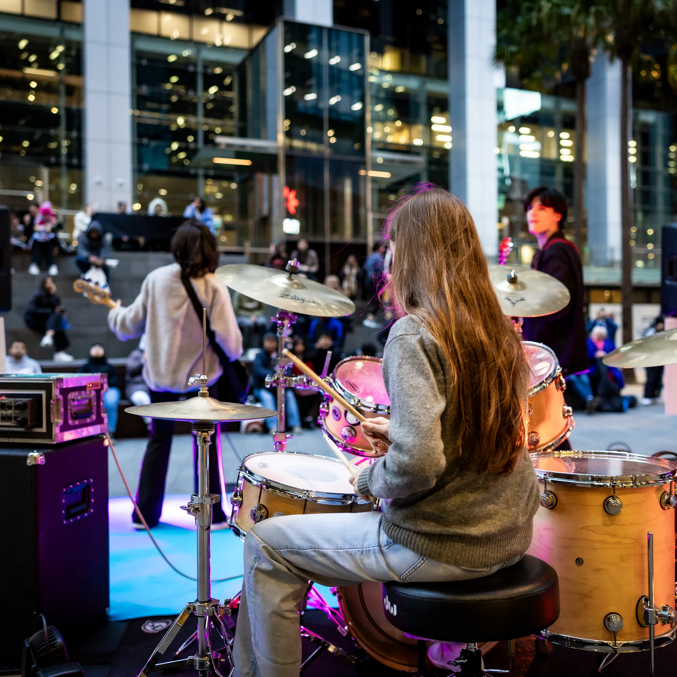 A long-haired woman plays a gold drum kit at a lively indoor concert, viewed from behind as the band and crowd gather beneath glass walls.
