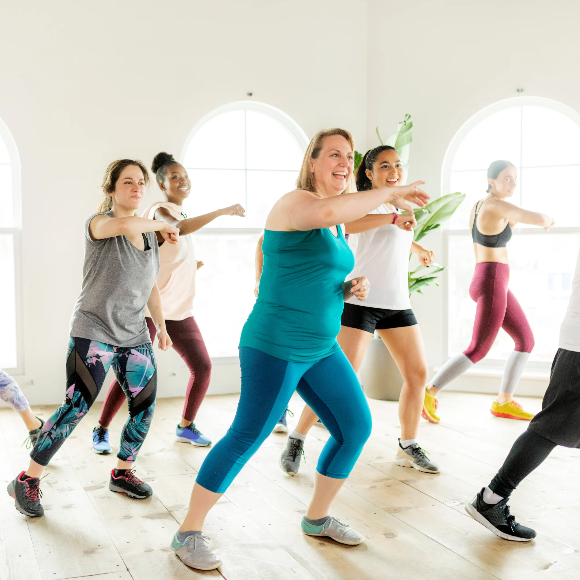 Adults in colourful workout clothes practice dance-fitness moves in a sunlit studio, following a woman in teal; the class feels upbeat and welcoming.