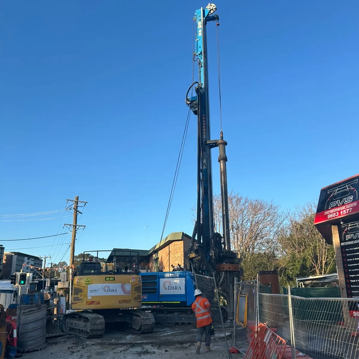 A towering blue drilling rig and excavators work at a fenced construction site beside a city street, with a worker in orange safety gear under a clear sky.