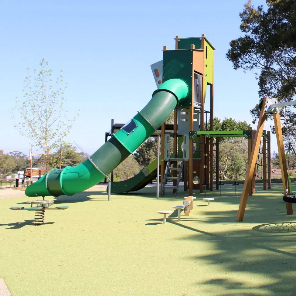 A sunny playground features a tall green-and-yellow climbing tower with a winding enclosed tube slide, swings, and spring riders on soft green ground.