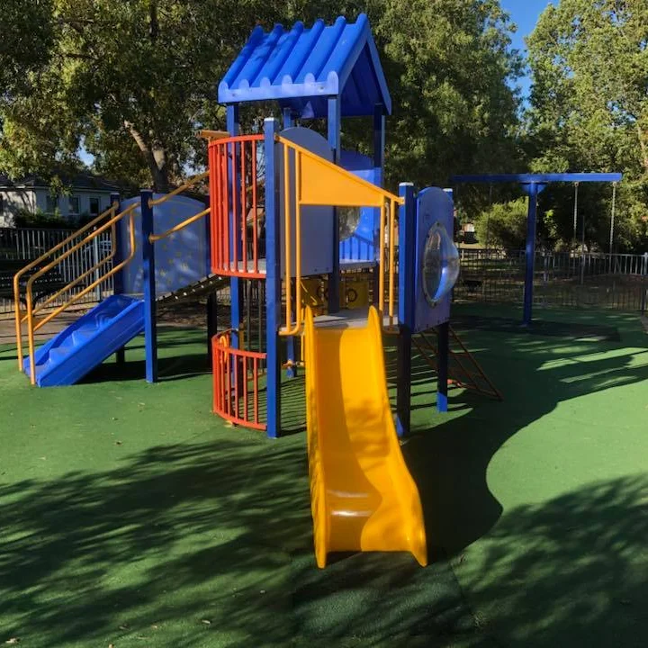 Colorful outdoor playground with blue and orange climbing structures, slides, and swings on green safety turf, surrounded by trees in sunlight.