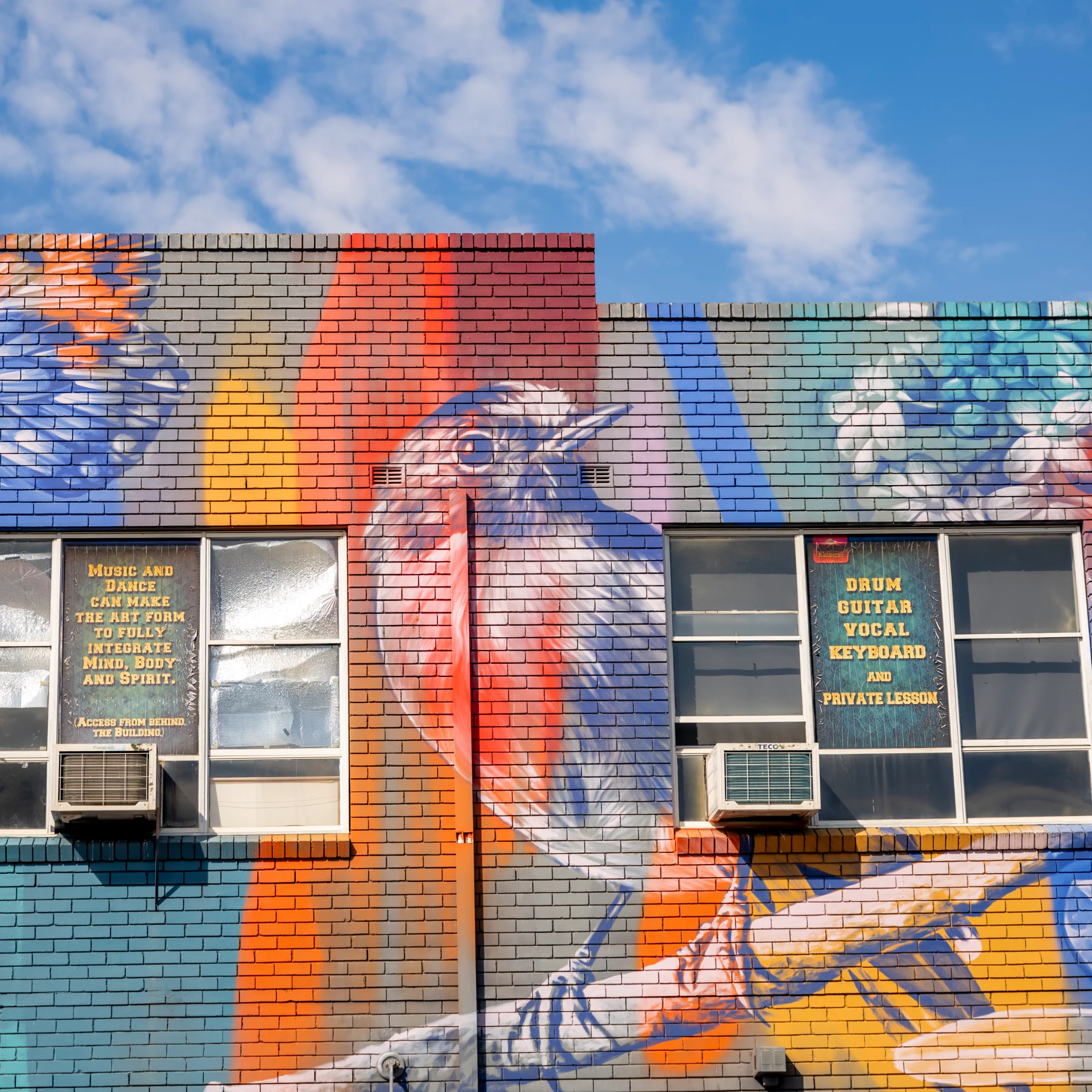 Vibrant urban mural on a brick building, featuring blue, orange, red, and yellow abstract shapes beneath a bright blue, cloud-filled sky.