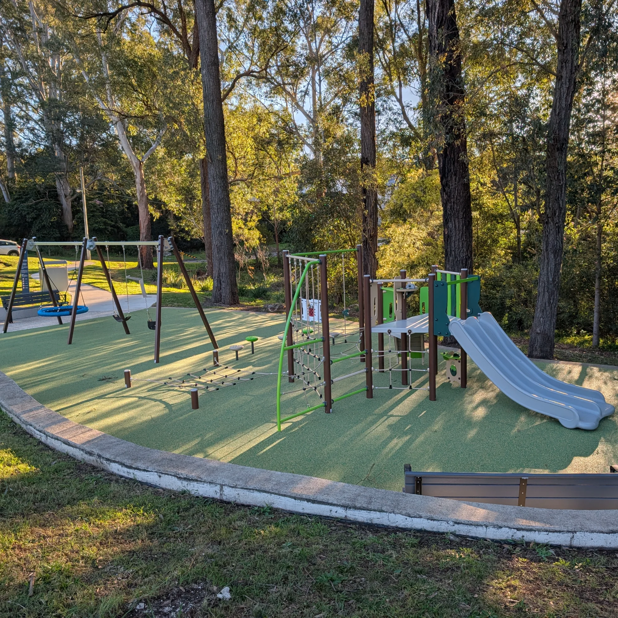 Sunlit playground with swings, climbing equipment, and a gray slide on green safety surfacing, nestled among tall trees in a peaceful park.