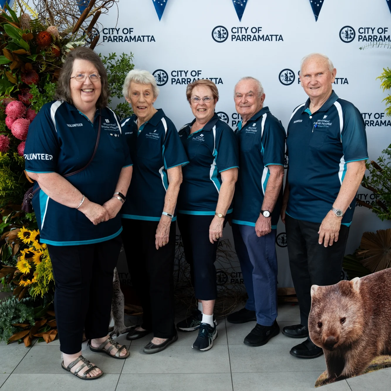 Five City of Parramatta volunteers pose in matching navy shirts beside vibrant floral displays, with a koala statue in the foreground.