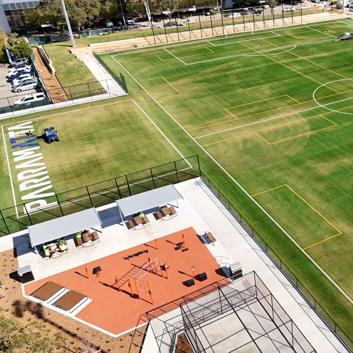 Aerial view of a sports complex with green soccer fields, an orange basketball court, playground, fencing, and surrounding urban buildings.