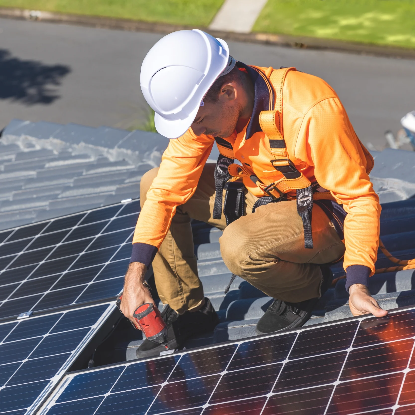 Solar technician in orange safety gear and hard hat installs rooftop photovoltaic panels, tightening a connection in bright sunlight.