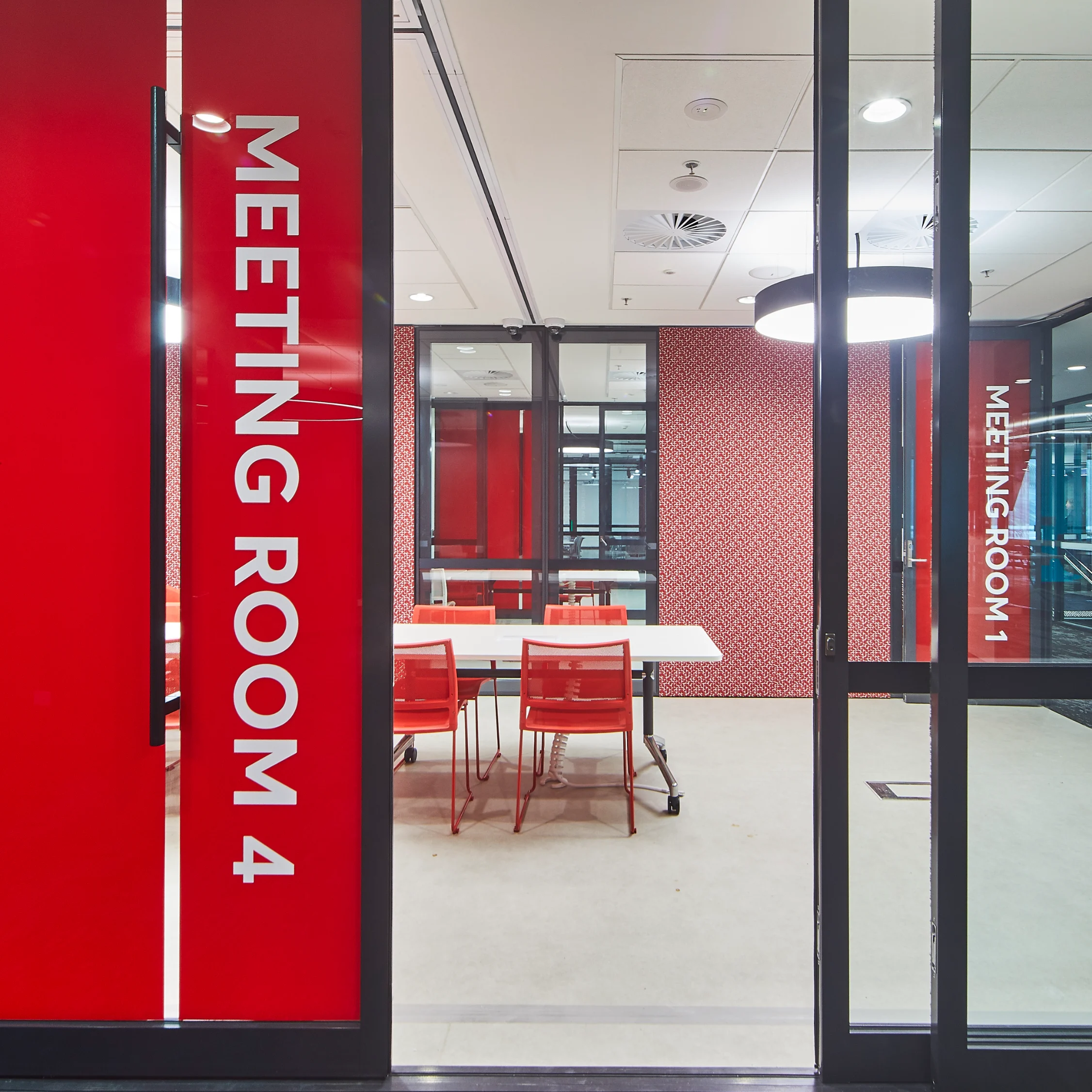 Modern glass-walled meeting room with bold red doors, white “Meeting Room 4” lettering, conference table, chairs, and bright interiors.