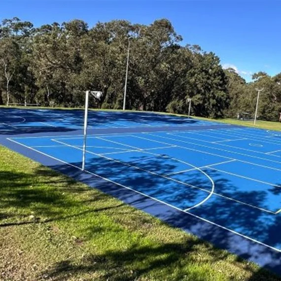 Empty blue outdoor tennis courts with white lines and nets, surrounded by green grass and trees under a clear blue sky; peaceful setting.