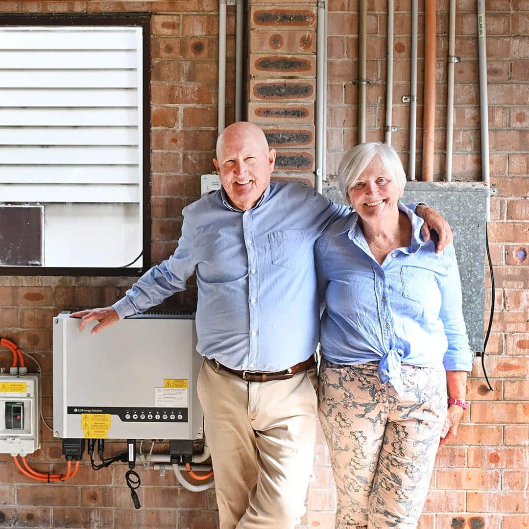 Smiling older couple stands beside a home solar battery and inverter system against a brick wall, conveying warmth, comfort, and clean energy.