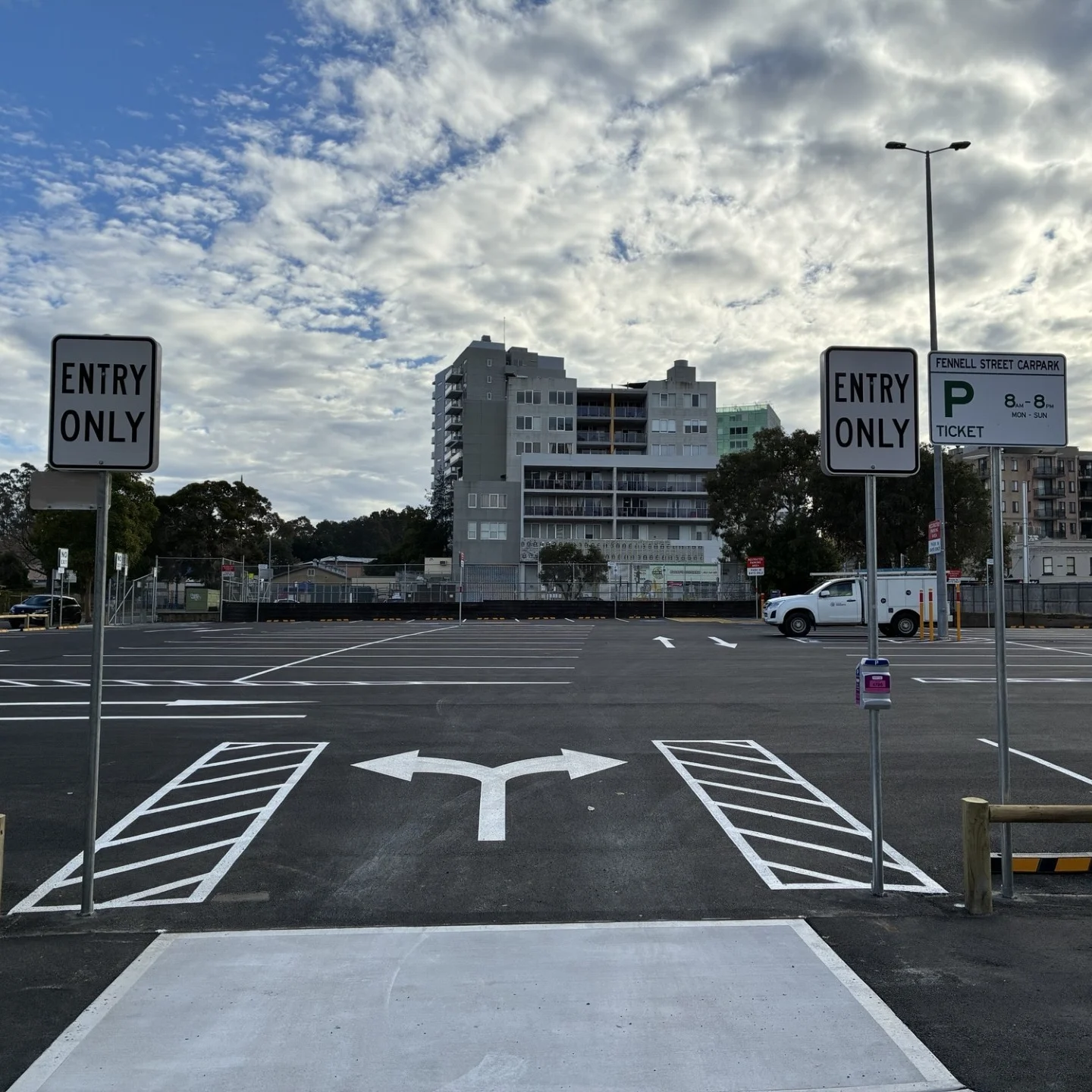 Nearly empty parking lot with entry-only signs, directional arrows, parking bays, city buildings, trees, and a dramatic cloudy sky.